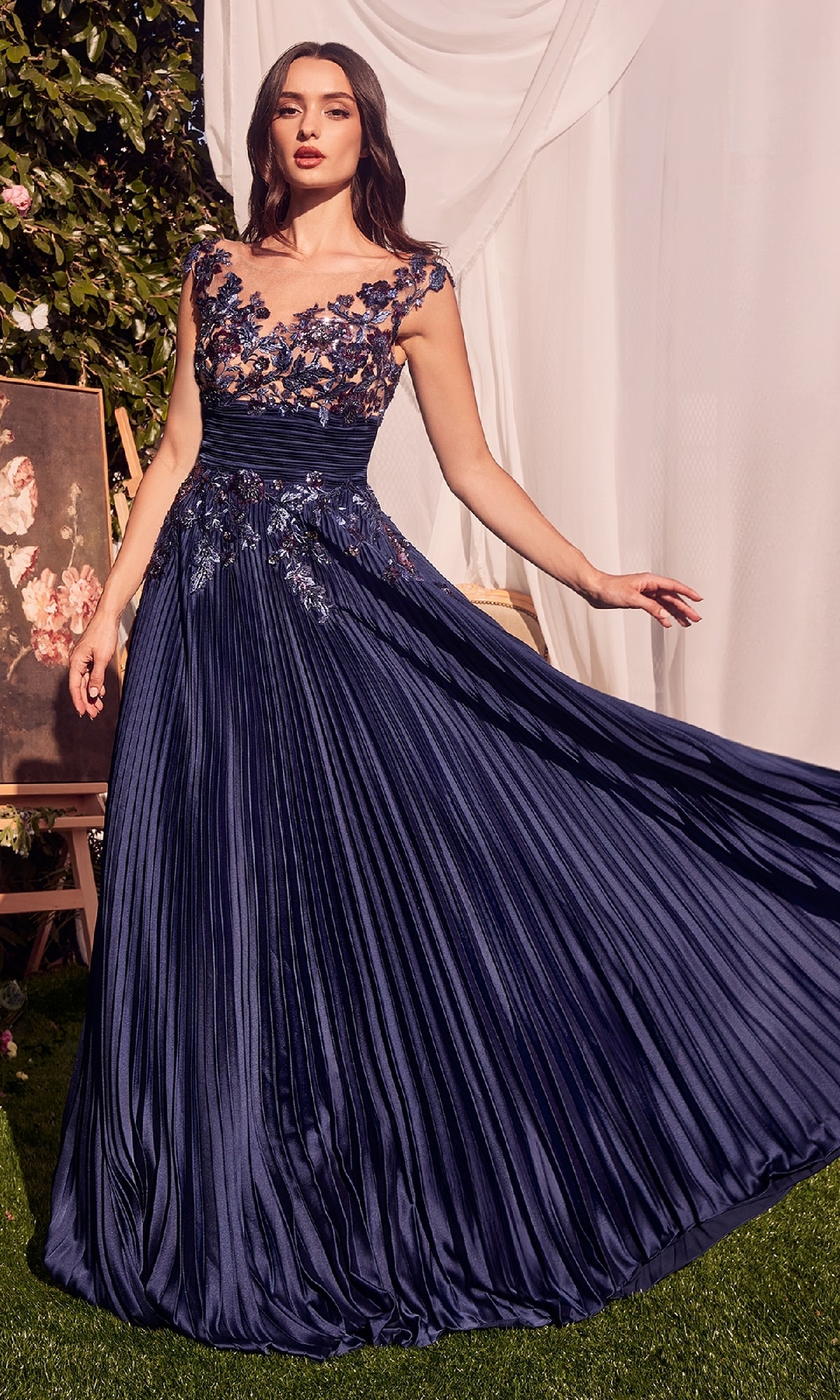 A woman poses outdoors in the Andrea & Leo A1355 Sequin Long Pleated Prom Dress, featuring sequin floral appliques on the bodice and shoulders, floor-length A-line silhouette, and extends one arm gracefully.